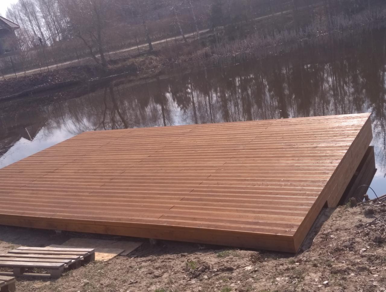 Nowo wybudowany pomost z desek kompozytowych, częściowo zanurzony w wodzie jeziora, z paletami budowlanymi leżącymi w pobliżu na brzegu.