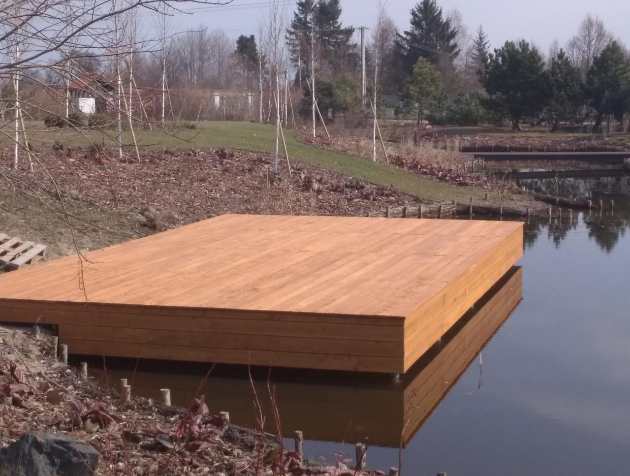 Drewniana platforma nad wodą, odbicie w tafli jeziora, widok z bliska, w tle drzewa i fragment budynku.