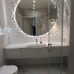 Gallen Sp Zoo - Nowoczesna łazienka z podświetlanym okrągłym lustrem, umywalką nablatową i szklaną kabiną prysznicową, wykończona płytkami z geometrycznym wzorem 3D.