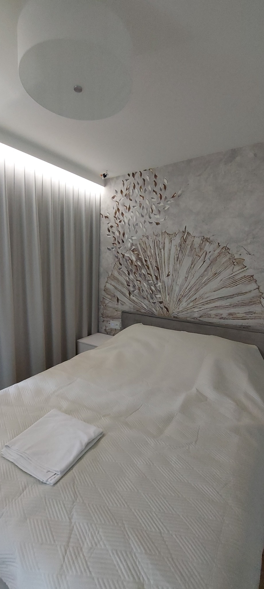 Sypialnia z łóżkiem przy ścianie z dekoracyjnym malunkiem w kształcie wachlarza i spadających liści, oświetlona lampą sufitową i bocznym światłem zza zasłon.