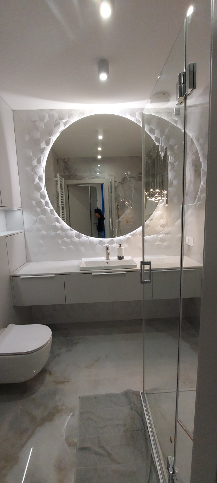 Nowoczesna łazienka z podświetlanym okrągłym lustrem, umywalką nablatową i szklaną kabiną prysznicową, wykończona płytkami z geometrycznym wzorem 3D.