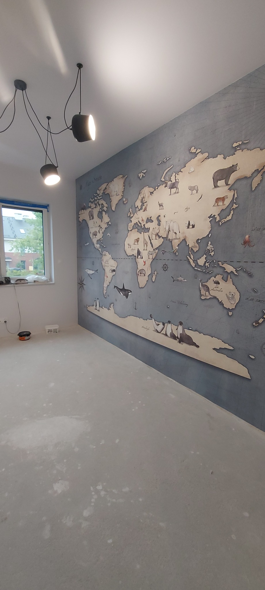 Ściana z fototapetą przedstawiającą mapę świata z wizerunkami zwierząt, nowoczesne oświetlenie sufitowe i surowy betonowy podkład podłogowy w pokoju przed wykończeniem.