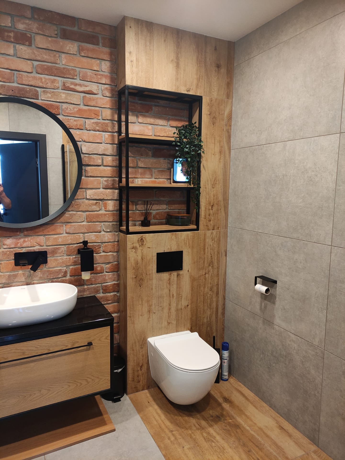 Nowoczesna łazienka z elementami cegły, drewnopodobnych płytek i szarości, okrągłe lustro z czarną ramą, wisząca toaleta i czarna armatura.
