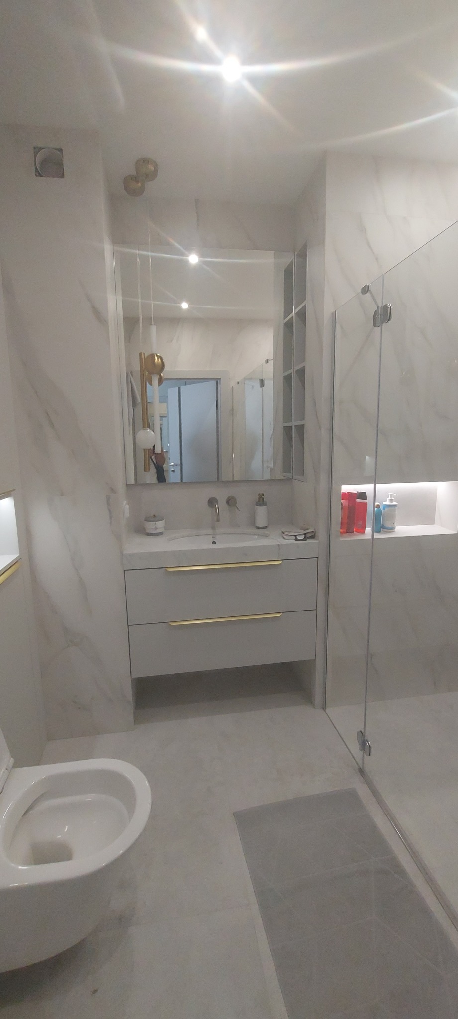 Nowoczesna łazienka z marmurowymi płytkami, szarą szafką pod umywalką z złotymi uchwytami, szklaną kabiną prysznicową i wiszącą lampą z elementami złota.