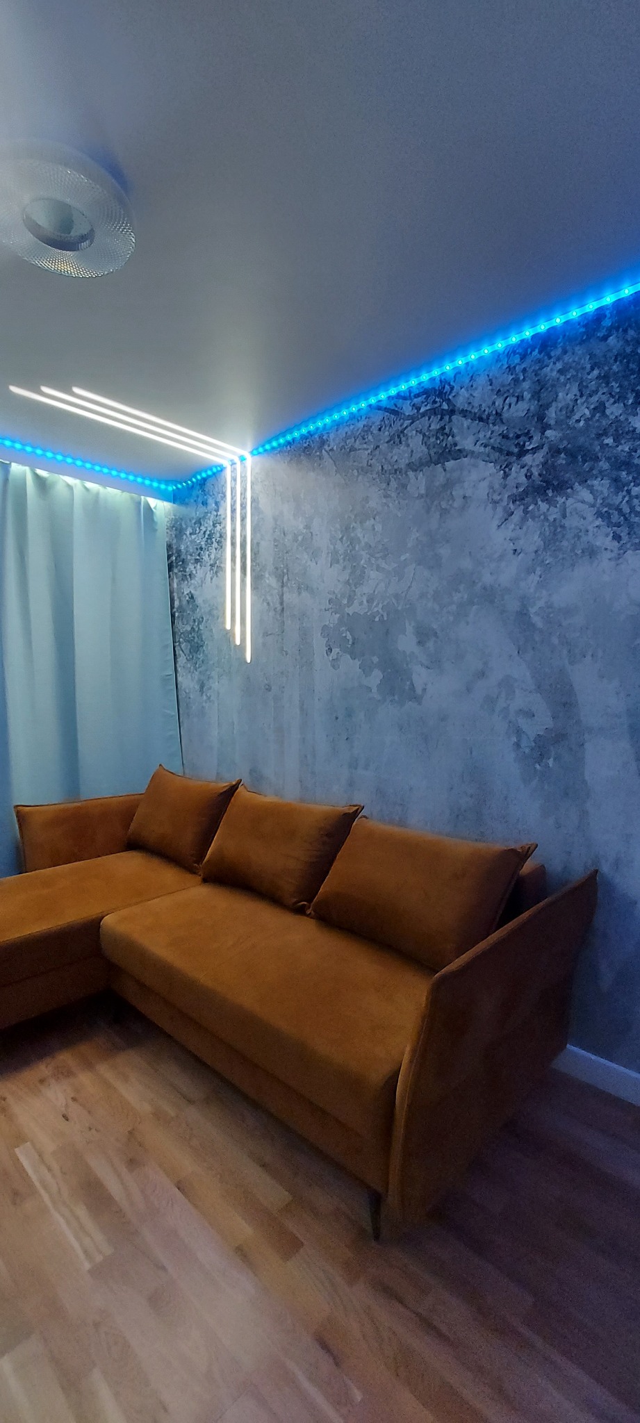 Brązowy narożnik z poduszkami w pokoju z dekoracyjną ścianą imitującą drzewo, oświetloną niebieskimi LED-ami i trzema pionowymi lampami.