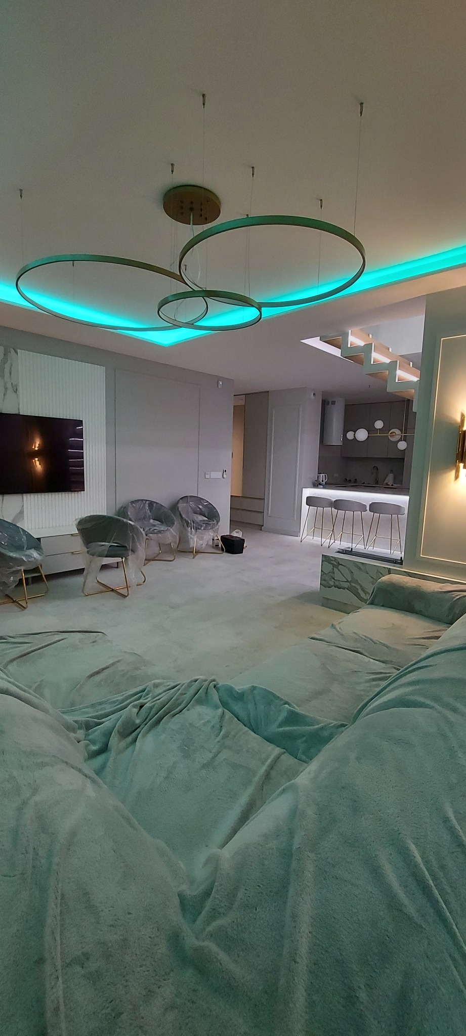 Wnętrze mieszkania w trakcie remontu z zakrytym folią meblem, widoczne krzesła zabezpieczone folią, nowoczesna lampa sufitowa z niebieskim podświetleniem LED i fragment kuchni z barkiem w tle.