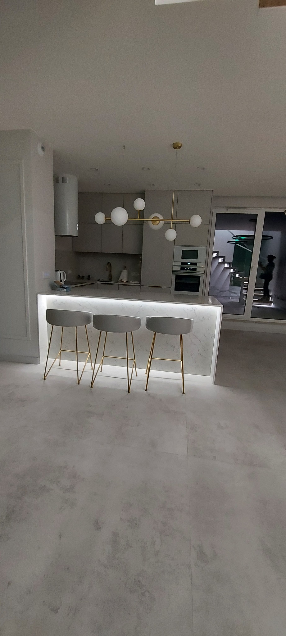 Nowoczesna kuchnia z wyspą wykończoną marmurem i podświetleniem LED, trzy szare hokery z złotymi nogami, minimalistyczna lampa sufitowa z kulistymi kloszami, zabudowa kuchenna w odcieniach szarości...