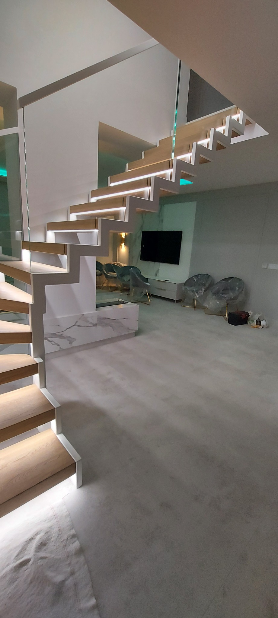 Nowoczesne schody dywanowe z podświetleniem LED, drewnianymi stopniami i szklaną balustradą w minimalistycznym wnętrzu, z widocznym salonem w tle.