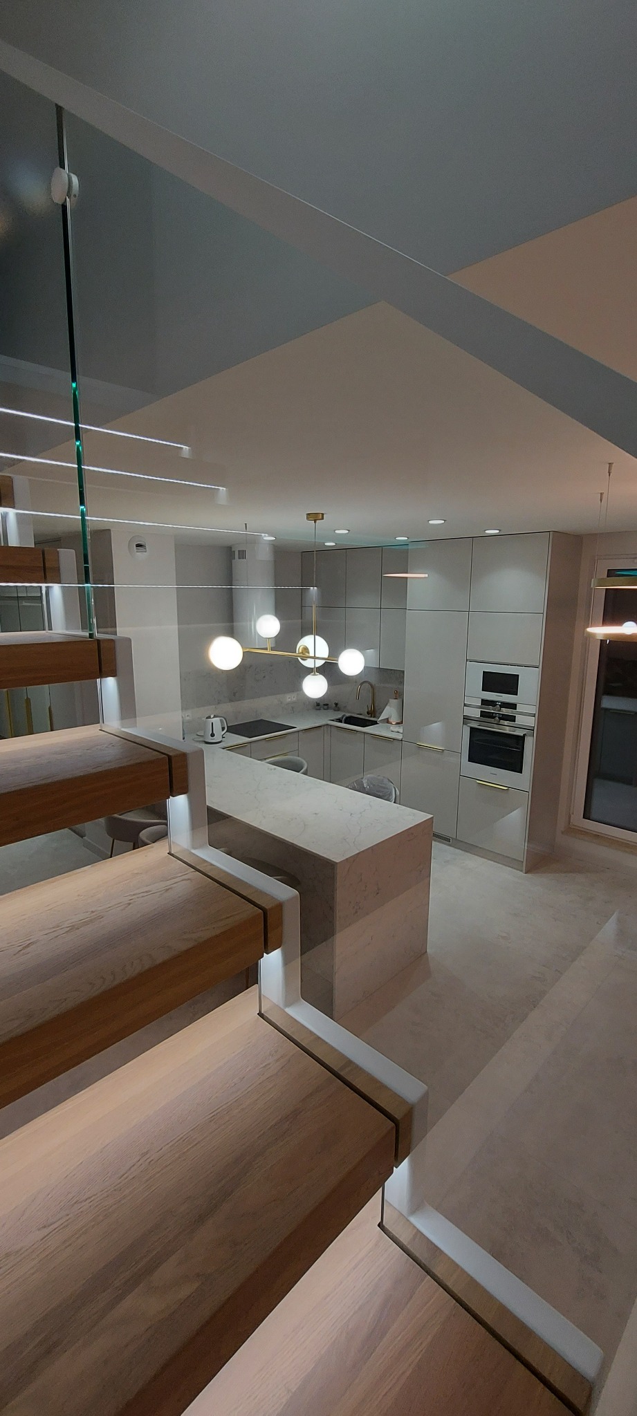Nowoczesna kuchnia z wyspą i oświetleniem kulistym, widziana z perspektywy drewnianych schodów ze szklaną balustradą.