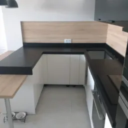 Nowoczesna kuchnia z czarnym blatem, jasnymi szafkami i drewnianym panelem ściennym, oświetlona wiszącymi lampami, z widocznym logo firmy w rogu.