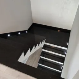 Czarne schody z kamienia z podświetleniem LED, widok z góry, białe akcenty na stopniach, logo firmy widoczne na ścianie.