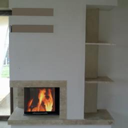 Floga - Nowoczesny kominek z obudową z jasnego kamienia, płomienie widoczne za szybą, obok półki wnękowe, logo firmy w prawym dolnym rogu.