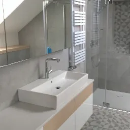 Nowoczesna łazienka na poddaszu z białą umywalką nablatową, szafką z drewnianymi akcentami, grzejnikiem drabinkowym i kabiną prysznicową z geometrycznym wzorem na ścianie, widoczny fragment lustra...
