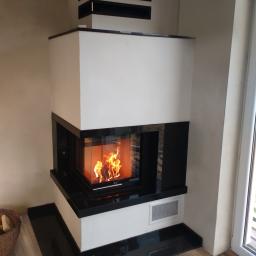 Floga - Nowoczesny kominek narożny z białym korpusem i czarnymi elementami wykończeniowymi, płomienie widoczne przez szklane drzwiczki, kosz z drewnem obok, logo firmy widoczne w rogu.