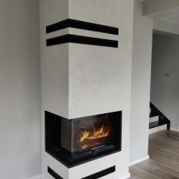 Floga - Biały narożny kominek z czarnymi, poziomymi akcentami, płomień widoczny przez szybę, na podłodze płytki imitujące drewno, w tle schody.