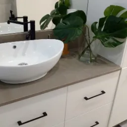 Biała, owalna umywalka nablatowa marki Massi na szarym blacie z konglomeratu, czarna bateria, monstera w szklanym wazonie, białe szafki z czarnymi uchwytami, logo firmy Flaga w prawym dolnym rogu.