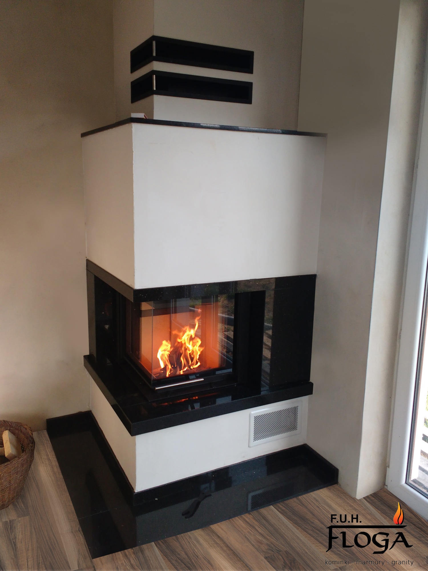 Nowoczesny kominek narożny z białym korpusem i czarnymi elementami wykończeniowymi, płomienie widoczne przez szklane drzwiczki, kosz z drewnem obok, logo firmy widoczne w rogu.