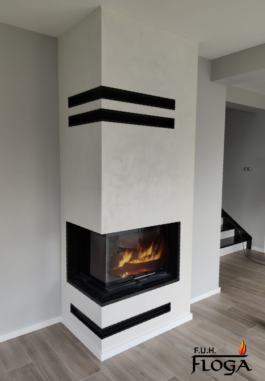 Biały narożny kominek z czarnymi, poziomymi akcentami, płomień widoczny przez szybę, na podłodze płytki imitujące drewno, w tle schody.