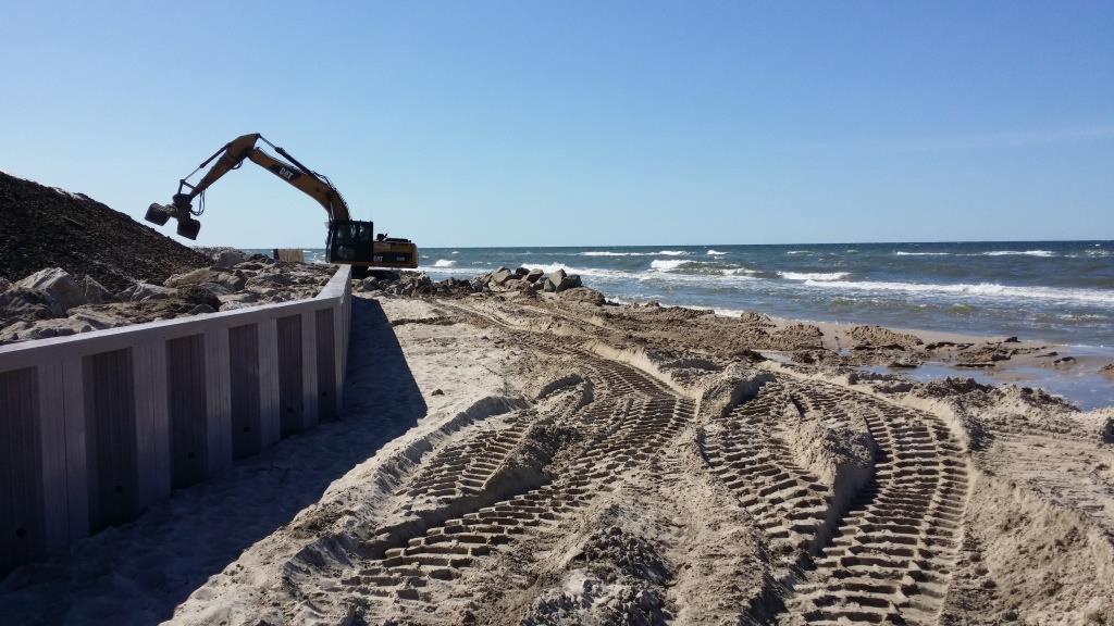 Brzeg morza z budowaną palisadą, piasek usiany śladami gąsienic koparki CAT układającej kamienie. W tle widoczne fale morskie i błękitne niebo.