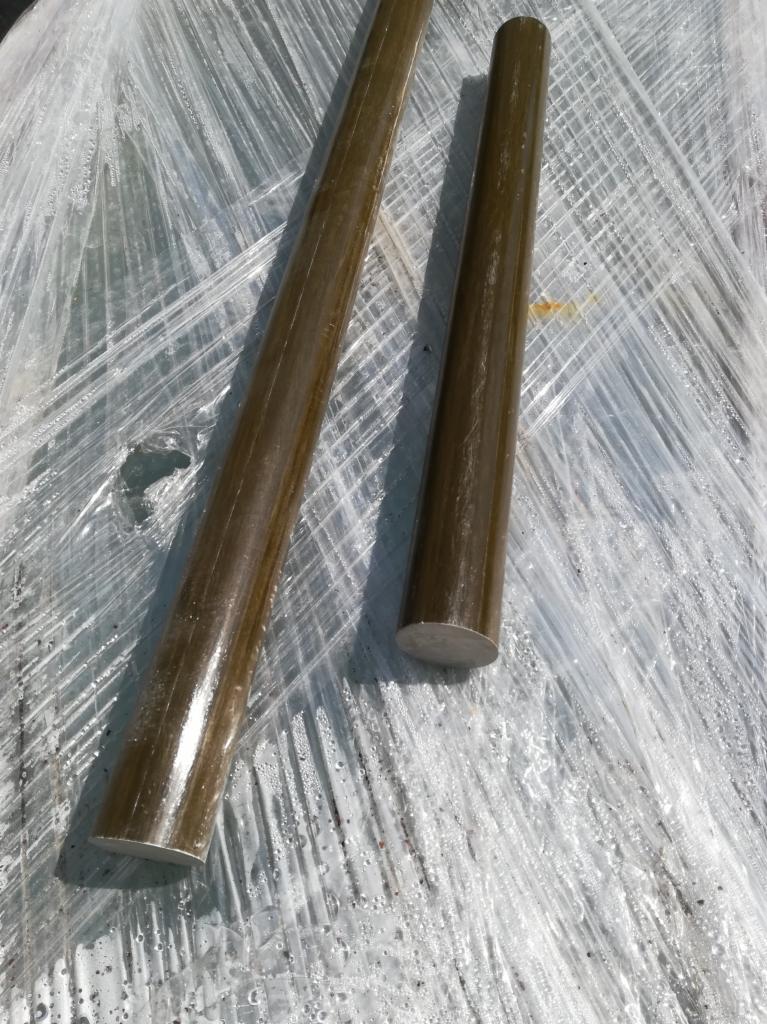 Dwa pręty zbrojeniowe o brązowym odcieniu, jeden długi i jeden krótki, leżą na folii ochronnej z widocznymi kroplami wody.