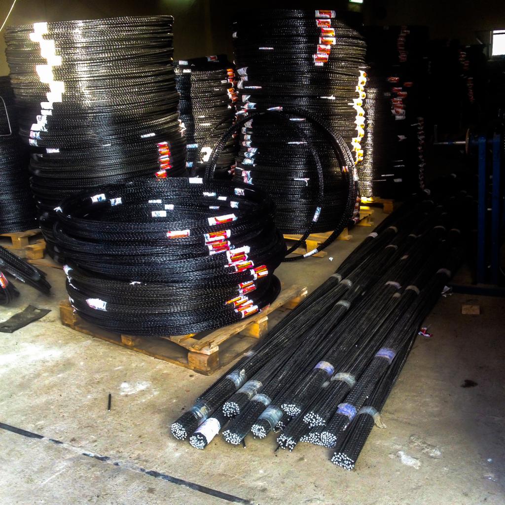 Magazyn z zapasem wiązek i kręgów czarnej stali, ułożonych na drewnianych paletach i bezpośrednio na betonowej posadzce.