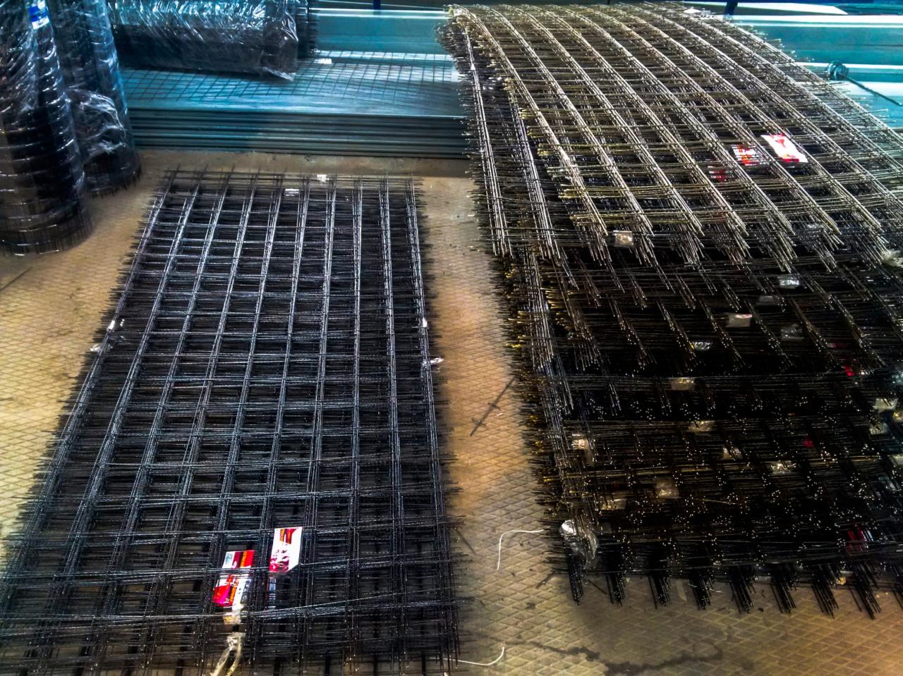 Siatki z drutu zbrojeniowego ułożone na betonowej posadzce; jedna rozłożona płasko, druga w stosie, widoczne etykiety producenta.