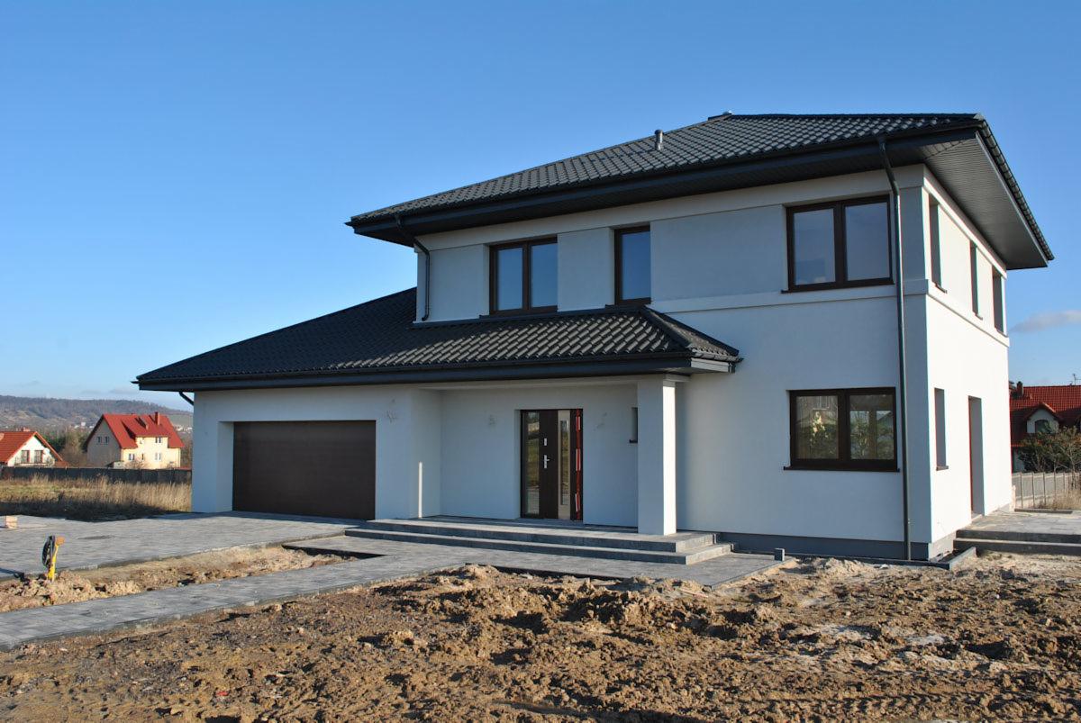 Nowoczesny dom dwukondygnacyjny z ciemną dachówką i brązowymi ramami okiennymi, w trakcie budowy chodnika z kostki brukowej na tle błękitnego nieba.