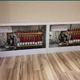 Dwa białe, zabudowane rozdzielacze ogrzewania podłogowego z czerwonymi i niebieskimi elementami, widoczne rury, pompy i okablowanie na tle drewnopodobnej podłogi.