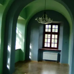 Wnętrze z łukowatymi przejściami pomalowane na zielono i niebiesko, z drewnianym oknem i grzejnikiem pod nim, widoczne refleksy światła słonecznego.
