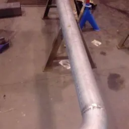 Długa, spawana rura ze stali nierdzewnej z kołnierzem, podparta na dwóch stojakach w warsztacie, widoczne ślady po spawaniu.