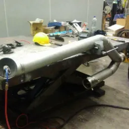 Metalowy rurociąg na warsztatowym stole z podnośnikiem, obok spawarka, szlifierka kątowa i żółty kask ochronny.