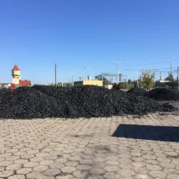 Góra czarnego węgla na betonowym placu z widoczną wieżą ciśnień w tle, oświetlona jasnym światłem słonecznym.
