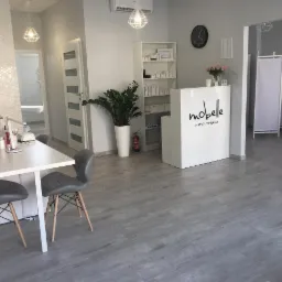 Jasne wnętrze salonu kosmetycznego w odcieniach szarości i bieli, z recepcją z logo 'mobelle Instytut Piękna', stołem do manicure z lampą i krzesłami, oraz dekoracyjną ścianą i roślinami doniczkowymi.