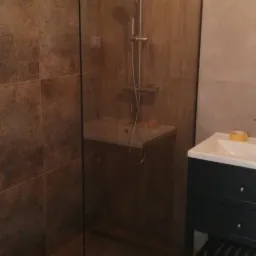 Nowoczesna kabina prysznicowa typu walk-in z brązowymi płytkami imitującymi drewno na ścianach i podłodze, biała umywalka nablatowa z szafką w kolorze granatowym.