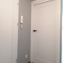 Dwa białe, wewnętrzne drzwi z poziomymi żłobieniami, ciemna klamka, szara ściana z domofonem i gniazdkiem elektrycznym, panele podłogowe w odcieniach brązu.