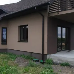 Wykończona elewacja domu jednorodzinnego z beżowym tynkiem, brązowymi oknami i balkonem, widoczne rury spustowe i trawnik.