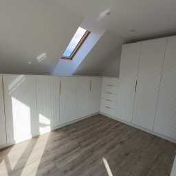 Red Tukan Barbara Jacaszek - Białe szafy na wymiar z ryflowanymi frontami i złotymi uchwytami, zamontowane na poddaszu ze skośnym sufitem i oknem dachowym. Jasna podłoga.