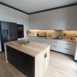 Nowoczesna kuchnia z wyspą: szare szafki z czarnymi uchwytami, drewniany blat, czarny sprzęt AGD i podświetlenie LED. Wyspa z czarnymi frontami.