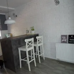 Kuchnia z blatem w kolorze grafitowym, dwoma białymi krzesłami barowymi z oparciami w kształcie litery X, białą ceglaną ścianą, grzejnikiem i drewnopodobną podłogą.