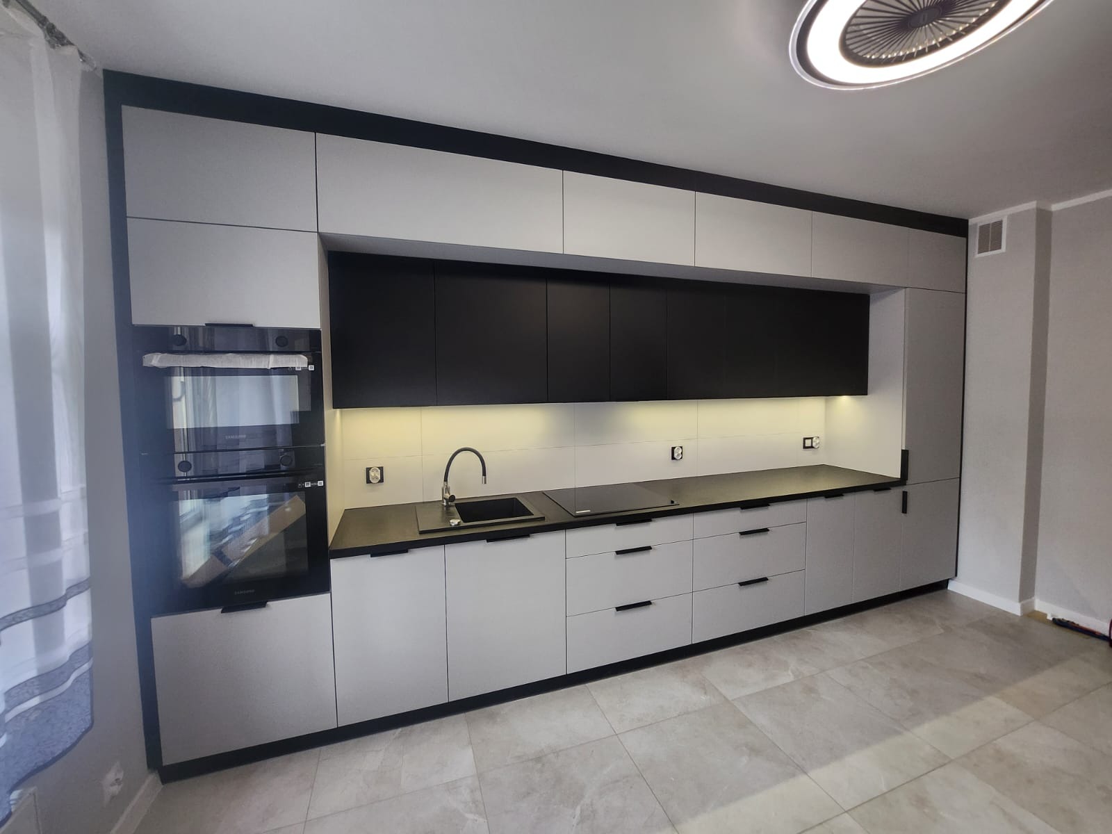 Nowoczesna kuchnia z szarymi szafkami dolnymi i czarnymi górnymi, oświetleniem LED podszafkowym i czarnym blatem. Uwagę zwraca designerska lampa sufitowa.