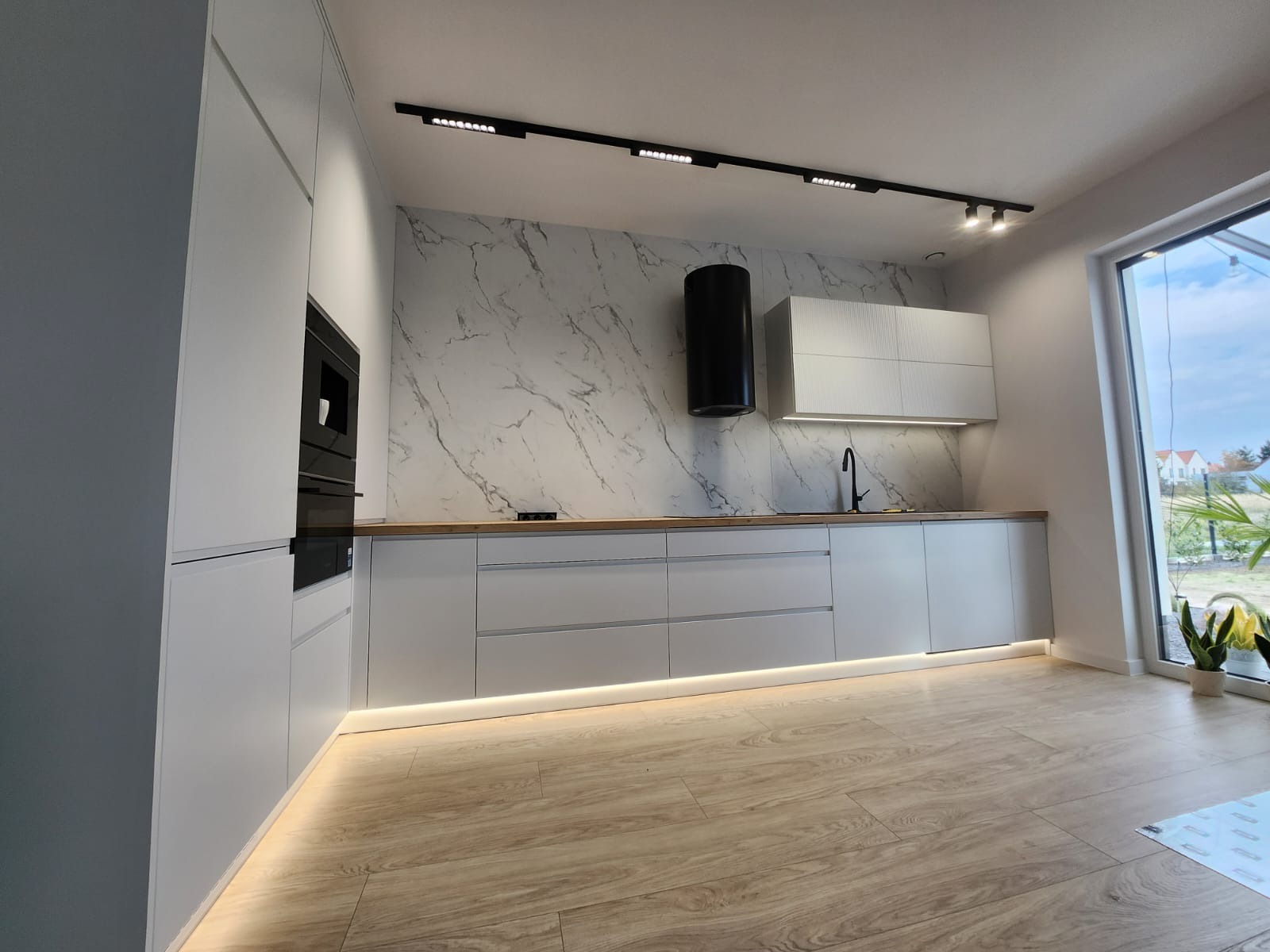 Nowoczesna kuchnia w Świdnicy z białymi frontami i marmurowym backsplashiem. Oświetlenie podszafkowe LED dodaje elegancji i podkreśla drewniany blat.