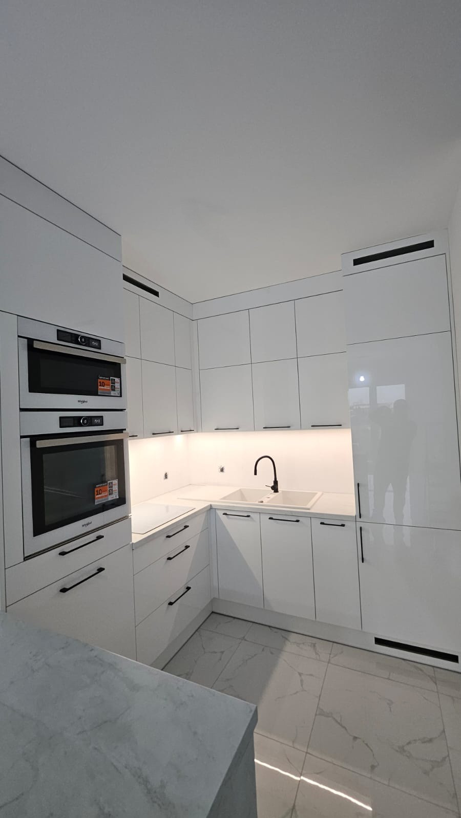Biała, nowoczesna kuchnia z połyskiem: zabudowa z piekarnikiem i mikrofalówką, czarne uchwyty, marmurowe płytki podłogowe, minimalistyczny design.