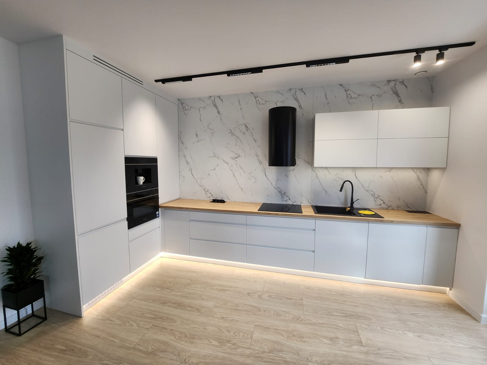 Nowoczesna, minimalistyczna kuchnia z białymi frontami, drewnianym blatem i marmurowym panelem ściennym. Oświetlenie podszafkowe LED i czarny okap cylindryczny.