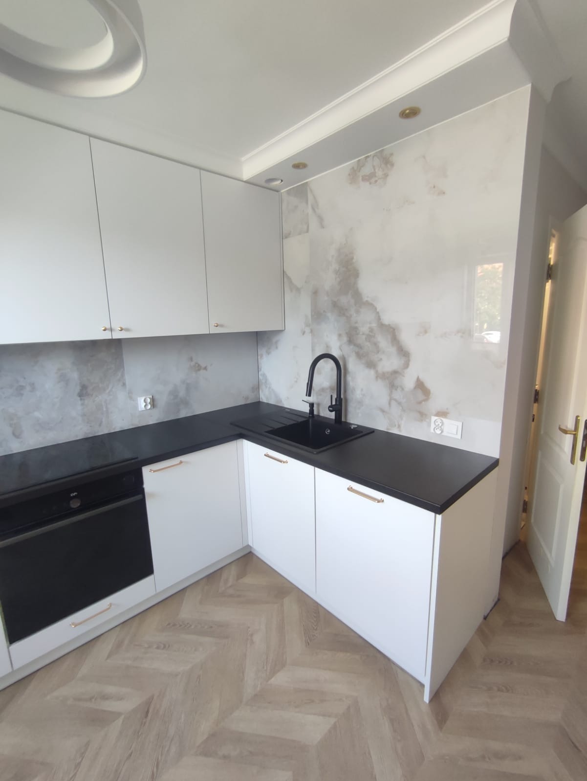 Biała kuchnia z czarnym blatem i armaturą, marmurowe płytki na ścianie, parkiet w jodełkę. Nowoczesne wykończenie, minimalistyczny styl, złote uchwyty.