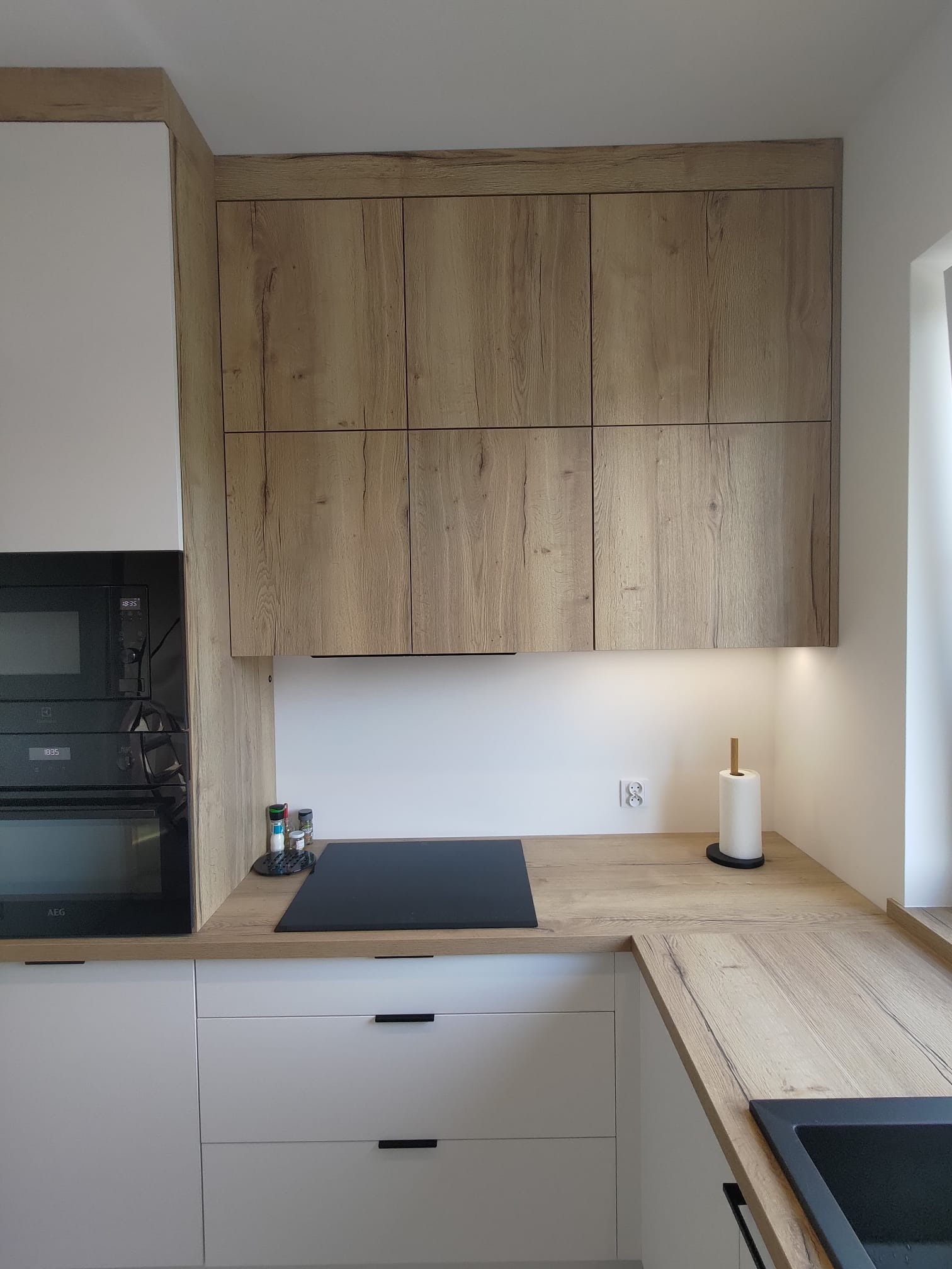 Jasna kuchnia z drewnianymi szafkami wiszącymi i białymi szafkami dolnymi. Widoczna płyta indukcyjna, piekarnik AEG i drewniany blat z akcesoriami.