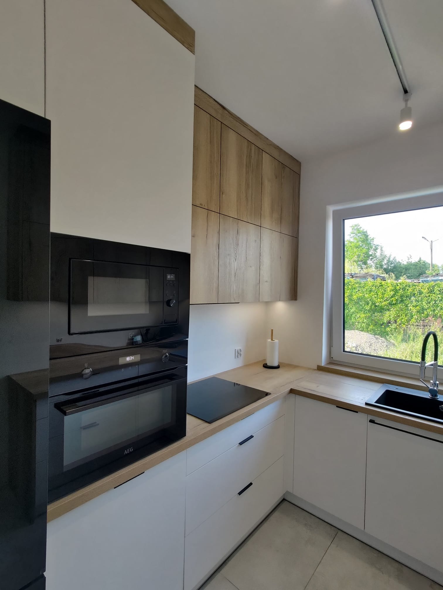 Nowoczesna kuchnia z zabudową w bieli i drewnie, czarne AGD (piekarnik, mikrofalówka, płyta) marki AEG. Blat drewniany, okno z widokiem na zieleń.