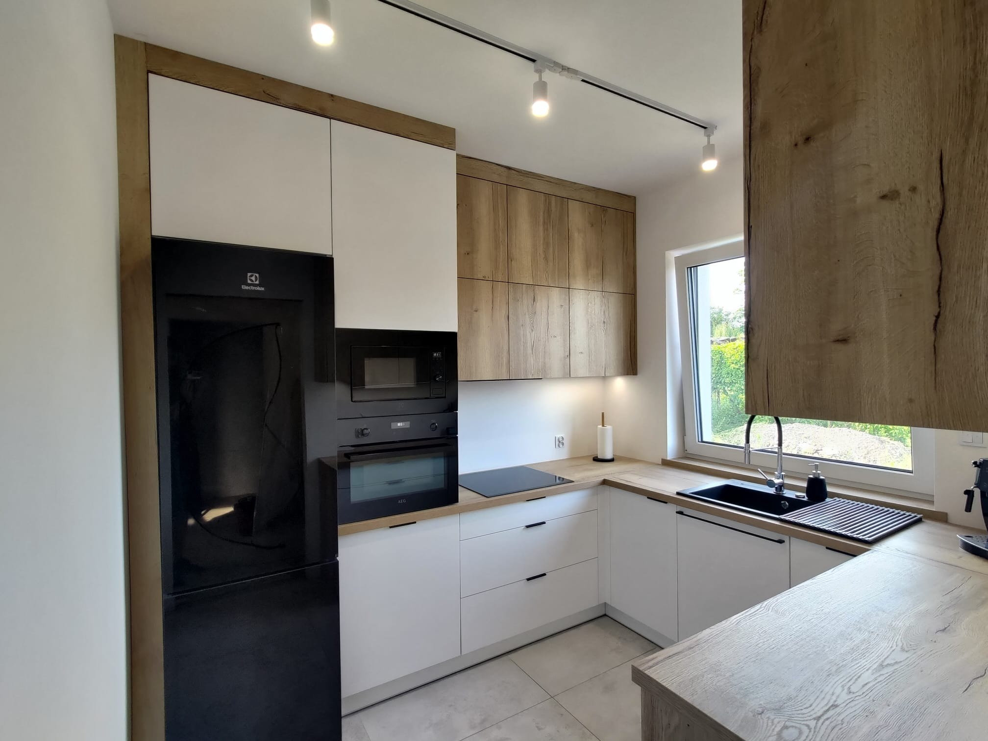 Nowoczesna kuchnia z białymi szafkami dolnymi i górnymi w odcieniu drewna, czarny sprzęt AGD, oświetlenie punktowe, jasna podłoga i blat.