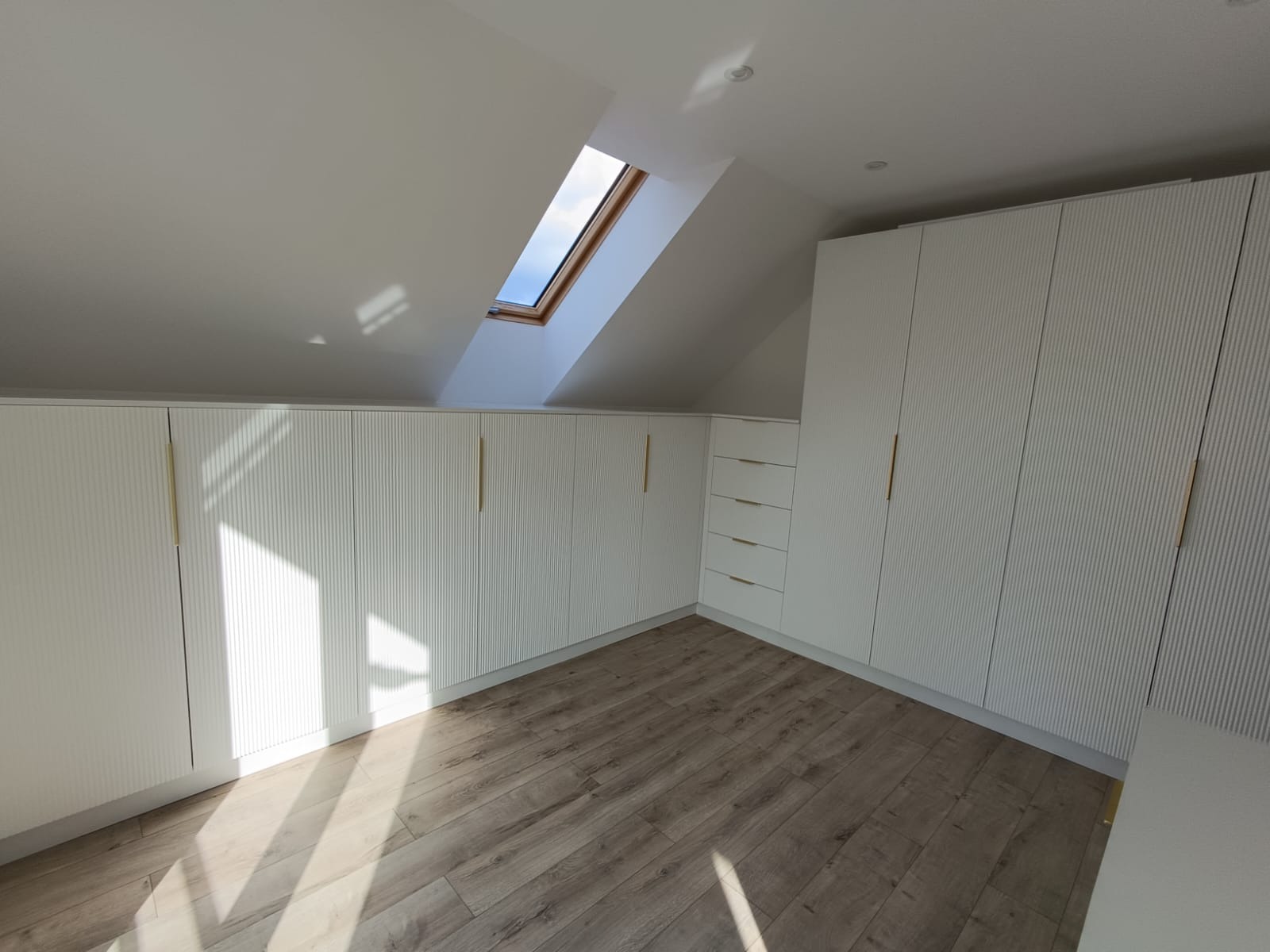 Białe szafy na wymiar z ryflowanymi frontami i złotymi uchwytami, zamontowane na poddaszu ze skośnym sufitem i oknem dachowym. Jasna podłoga.