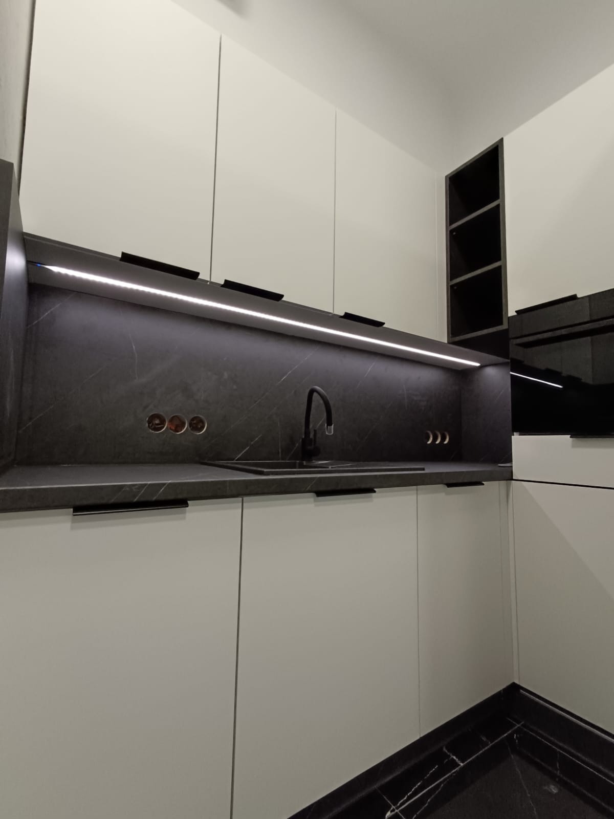 Nowoczesna kuchnia z białymi frontami, czarnym blatem i oświetleniem LED. Minimalistyczny design z czarną armaturą i marmurową podłogą. Widoczne gniazdka i półki.