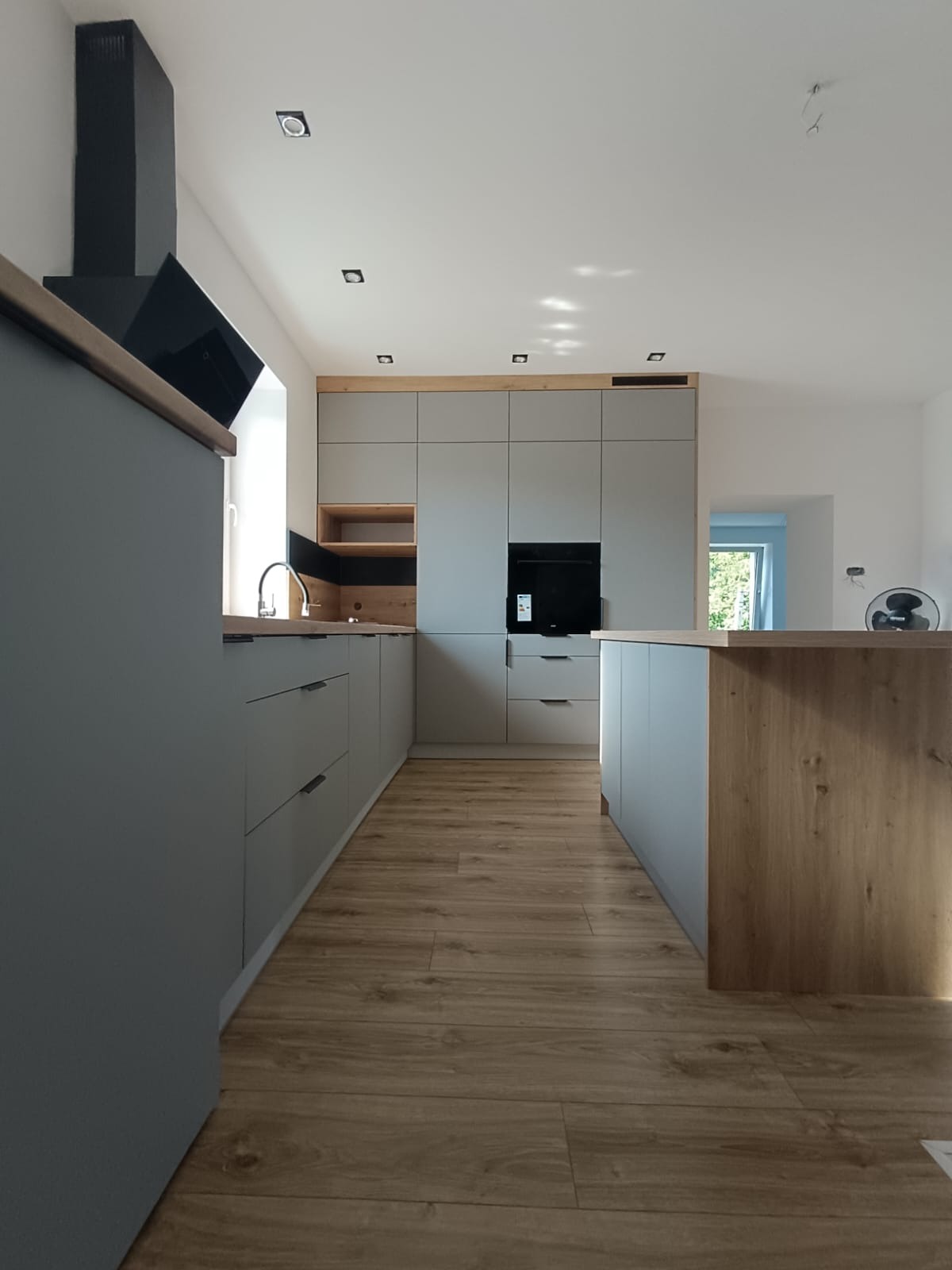 Szara kuchnia z drewnianymi akcentami: zabudowa meblowa, wyspa kuchenna, podłoga imitująca drewno. Minimalistyczny design z czarnym okapem.